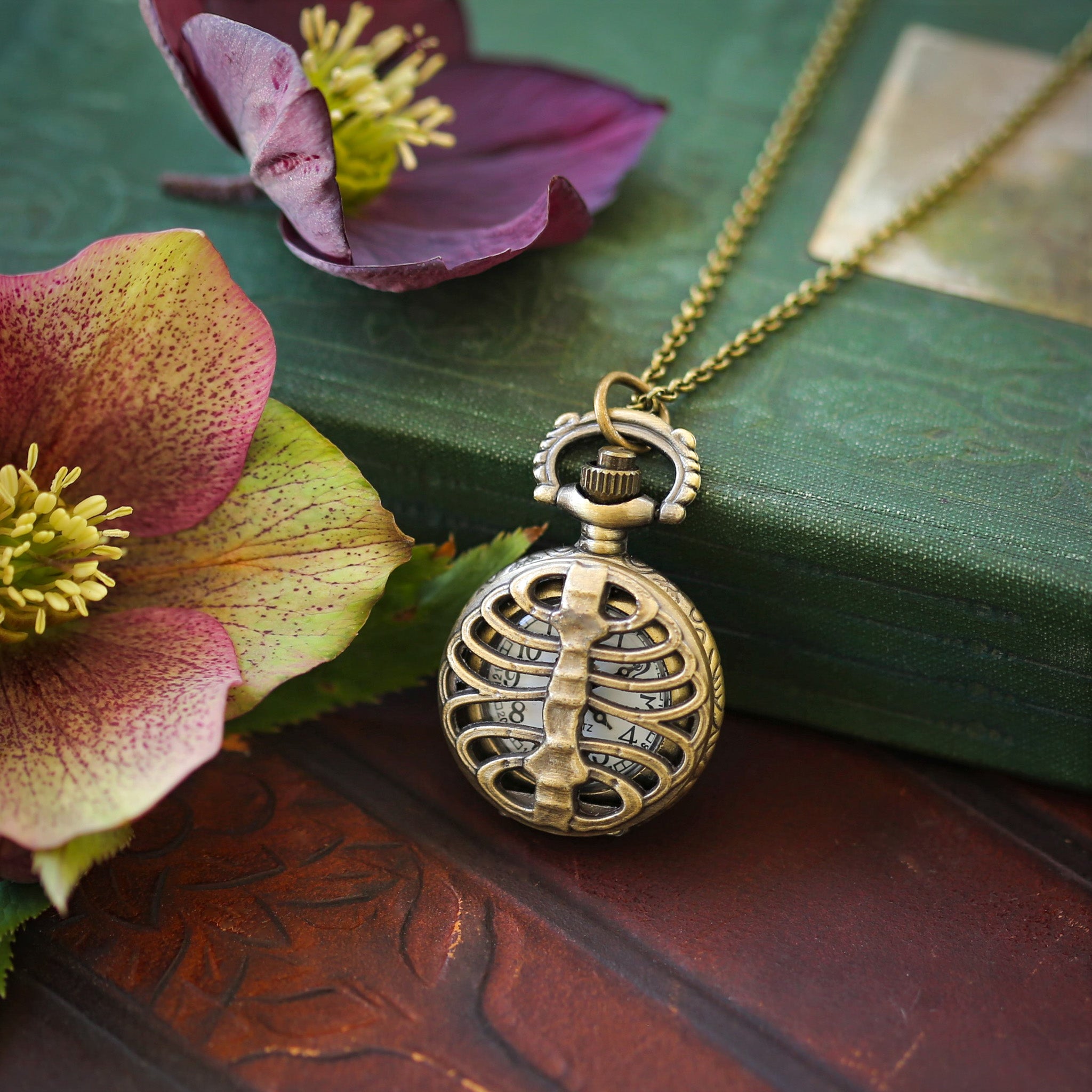 A brass-colored pocket watch necklace with an intricate ribcage design displayed on a book with a flower beside it.