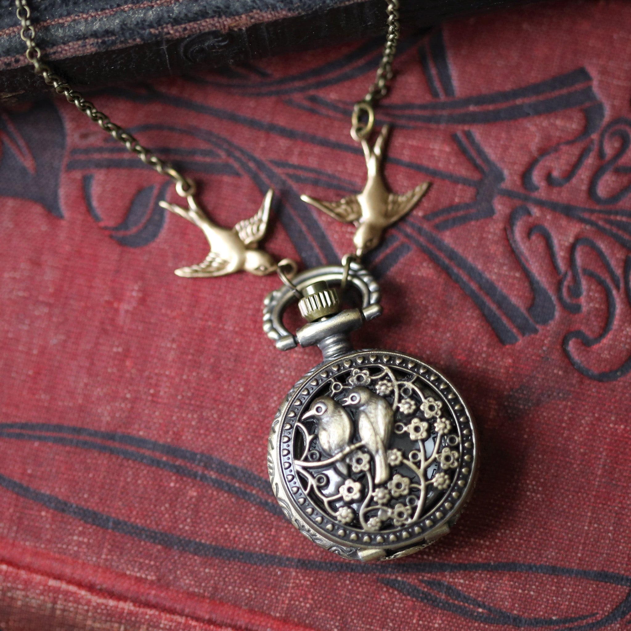 Small brass pocket watch with a cover design of two birds sitting on floral branches. Connecting the watch to the chain are two bird charms.