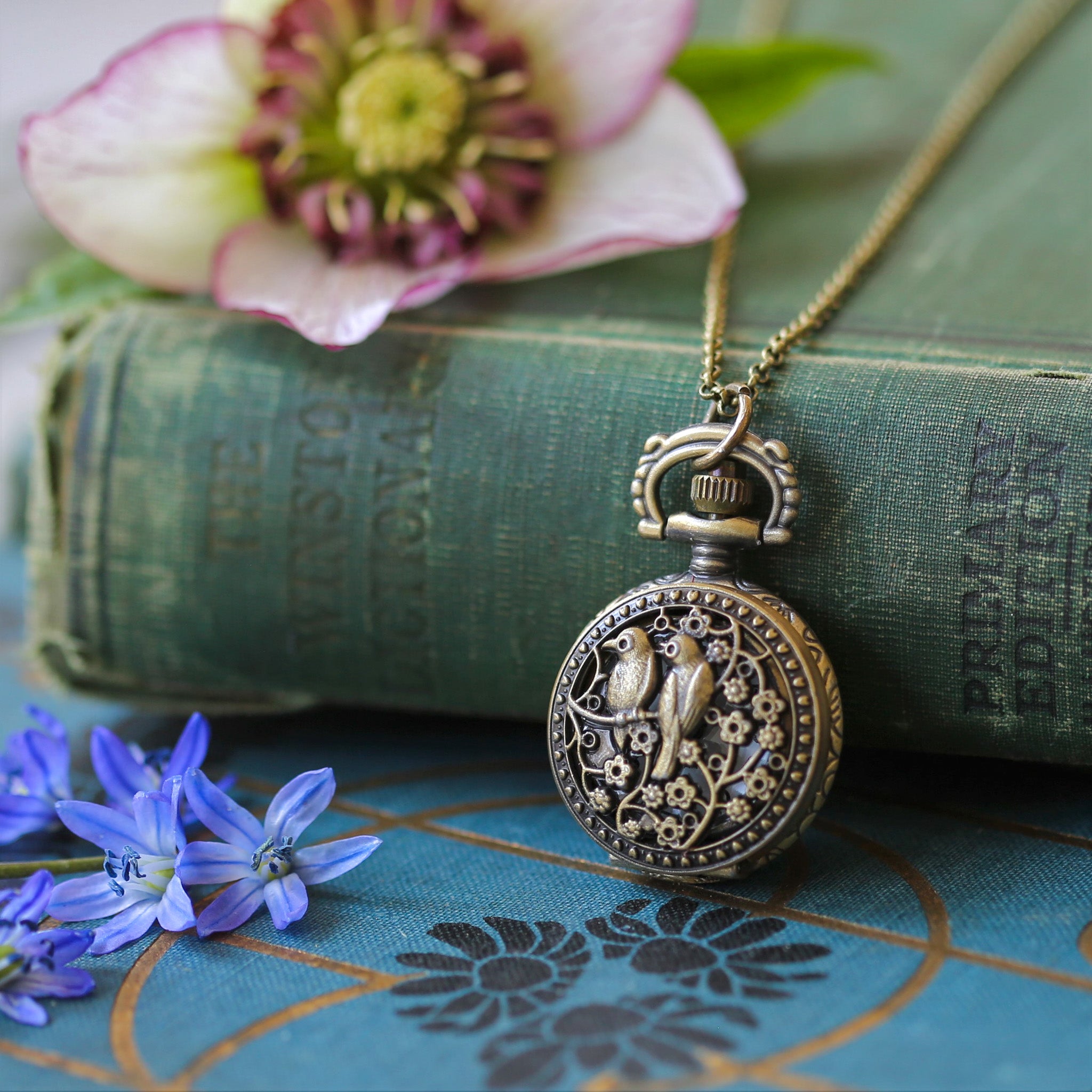 Small brass pocket watch with a cover design of two birds sitting on floral branches. Watch is on a simple brass chain.