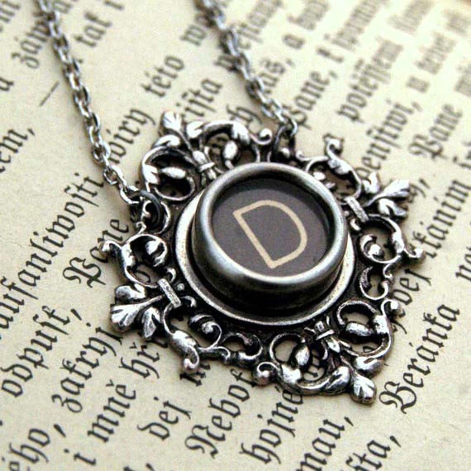 A typewriter key set on a decorative filigree pendant on a necklace chain. Typewriter key is black with an off-white colored letter D.