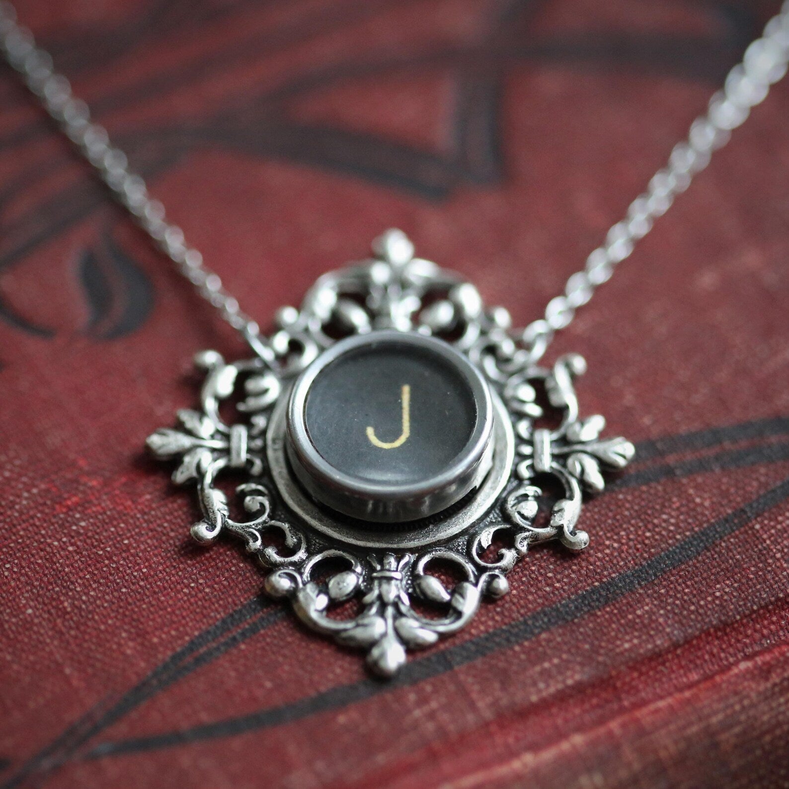 A typewriter key set on a decorative filigree pendant on a necklace chain. Typewriter key is black with an off-white colored letter J.