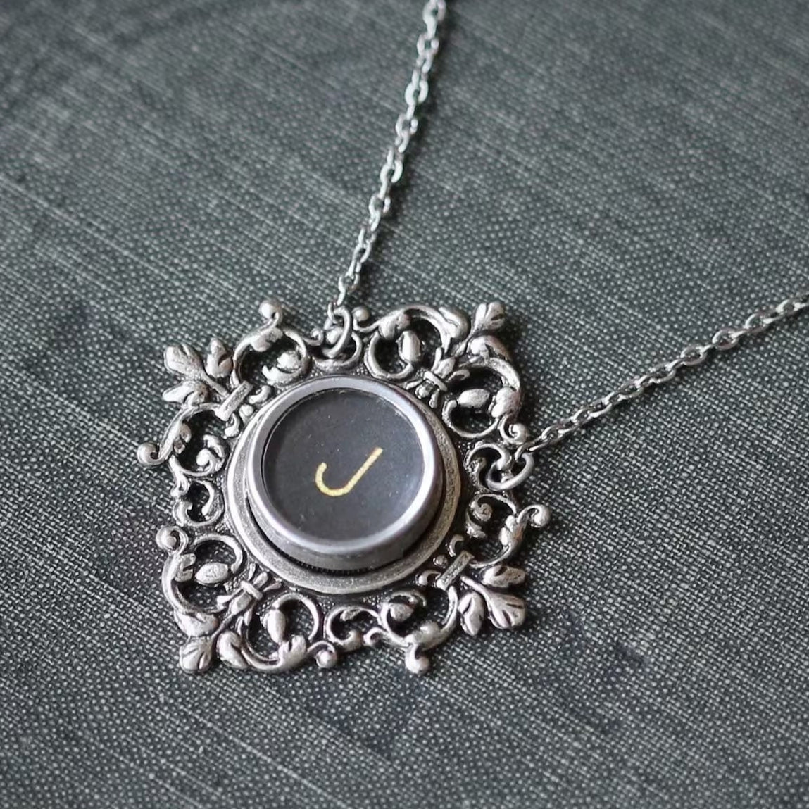 A typewriter key set on a decorative filigree pendant on a necklace chain. Typewriter key is black with a cream-colored letter J.