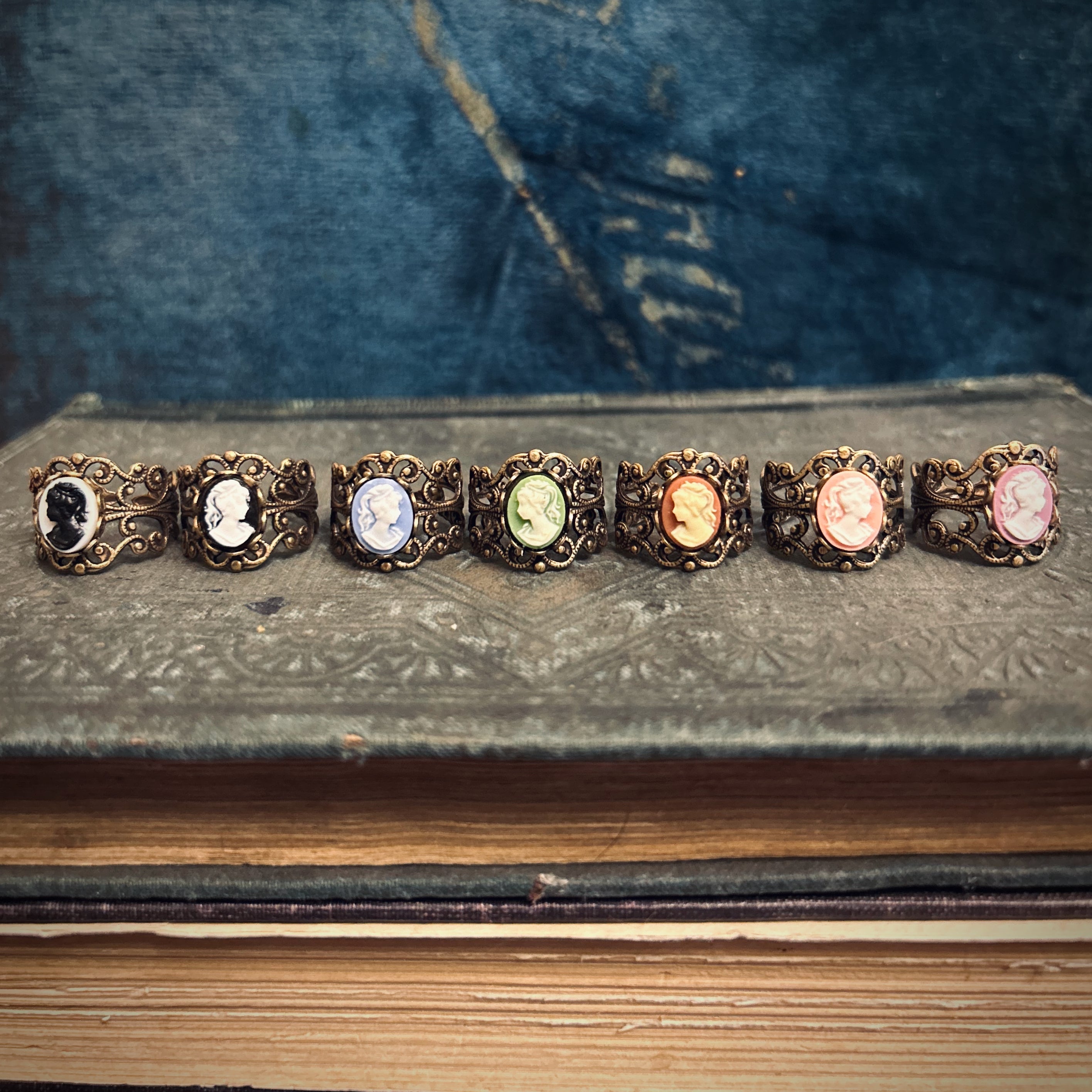 Seven brass adjustable filigree cameo rings with different colored profile cameos. The options are a black profile on white, white on black, cream on orange, white on green, white on blue, white on pink, and cream on peach.