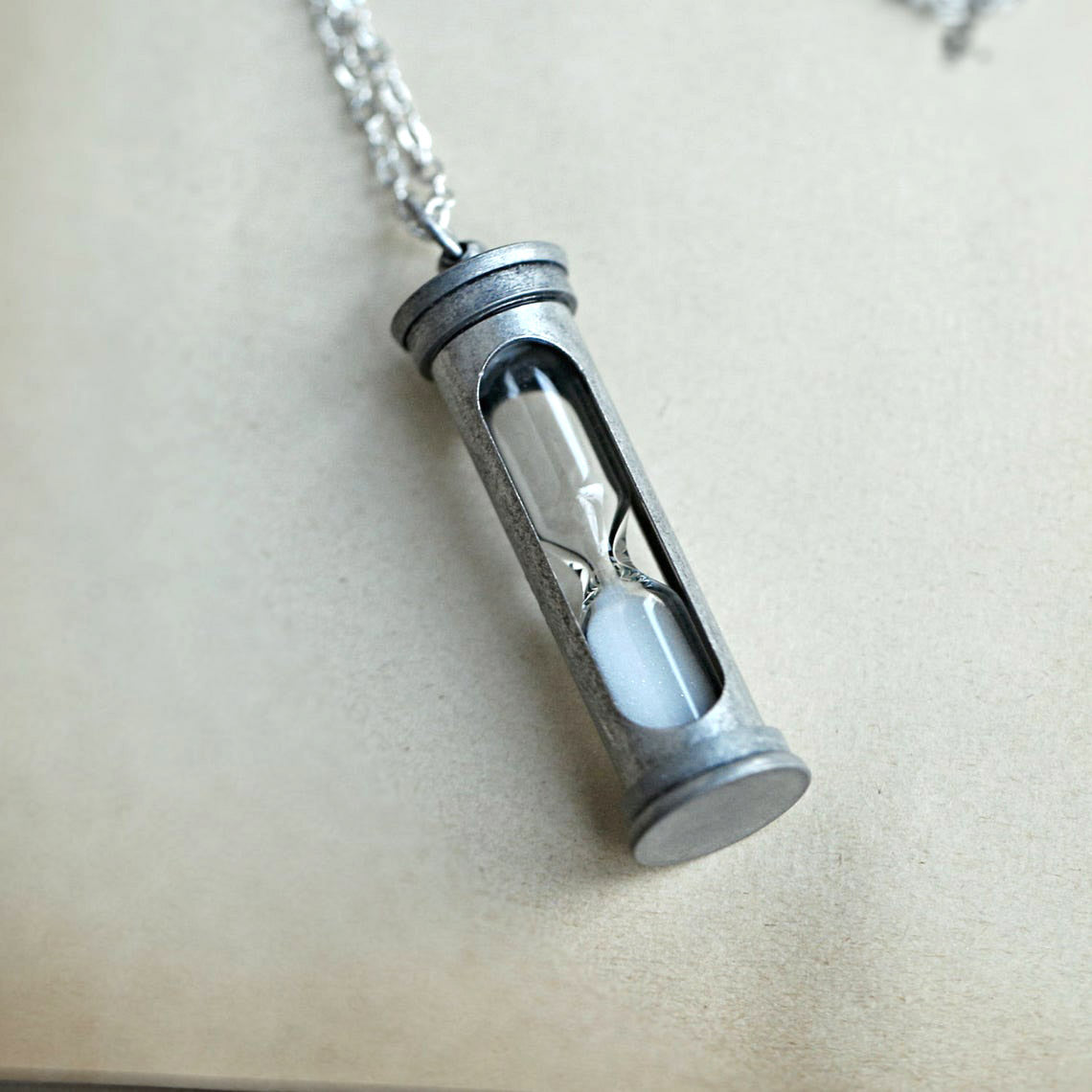 A functional hourglass in an antiqued silver frame hanging from a silver chain. The sand timer's sand is white.