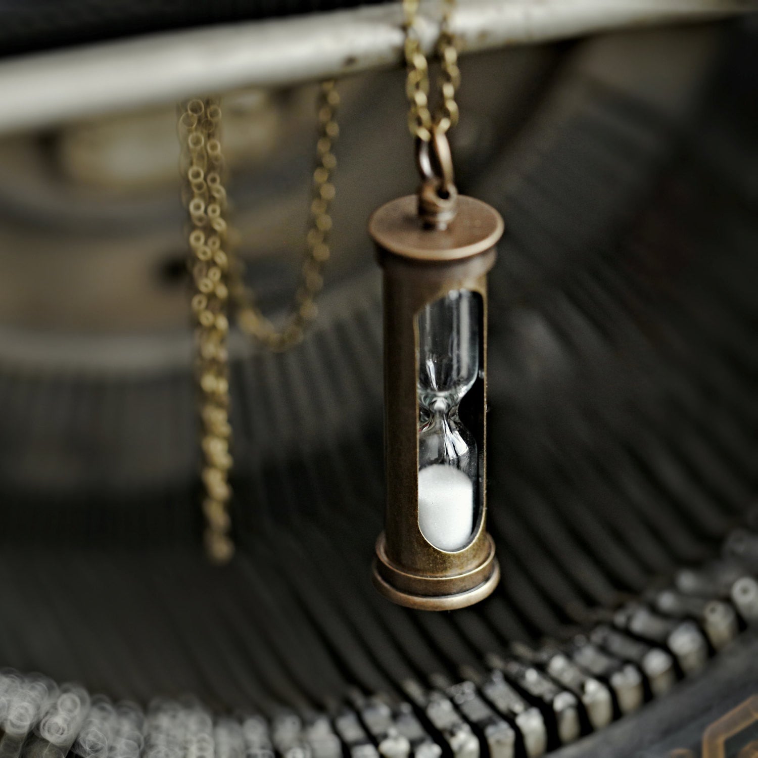A functional hourglass in an antiqued brass frame hanging from a brass chain. The sand timer's sand is white.