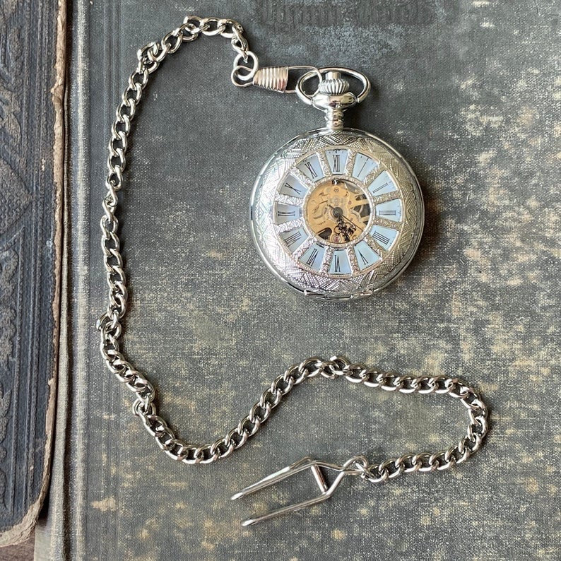 Silver mechanical pocket watch with a cogwheel design on the front, attached to a silver watch chain. The watch is closed, but internal gears and numerals are visible through the lid.