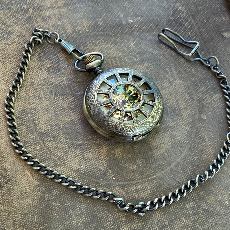 Antiqued brass mechanical pocket watch with a cogwheel design on the front, attached to a brass watch chain. The watch is closed, but internal gears and numerals are visible through the lid.