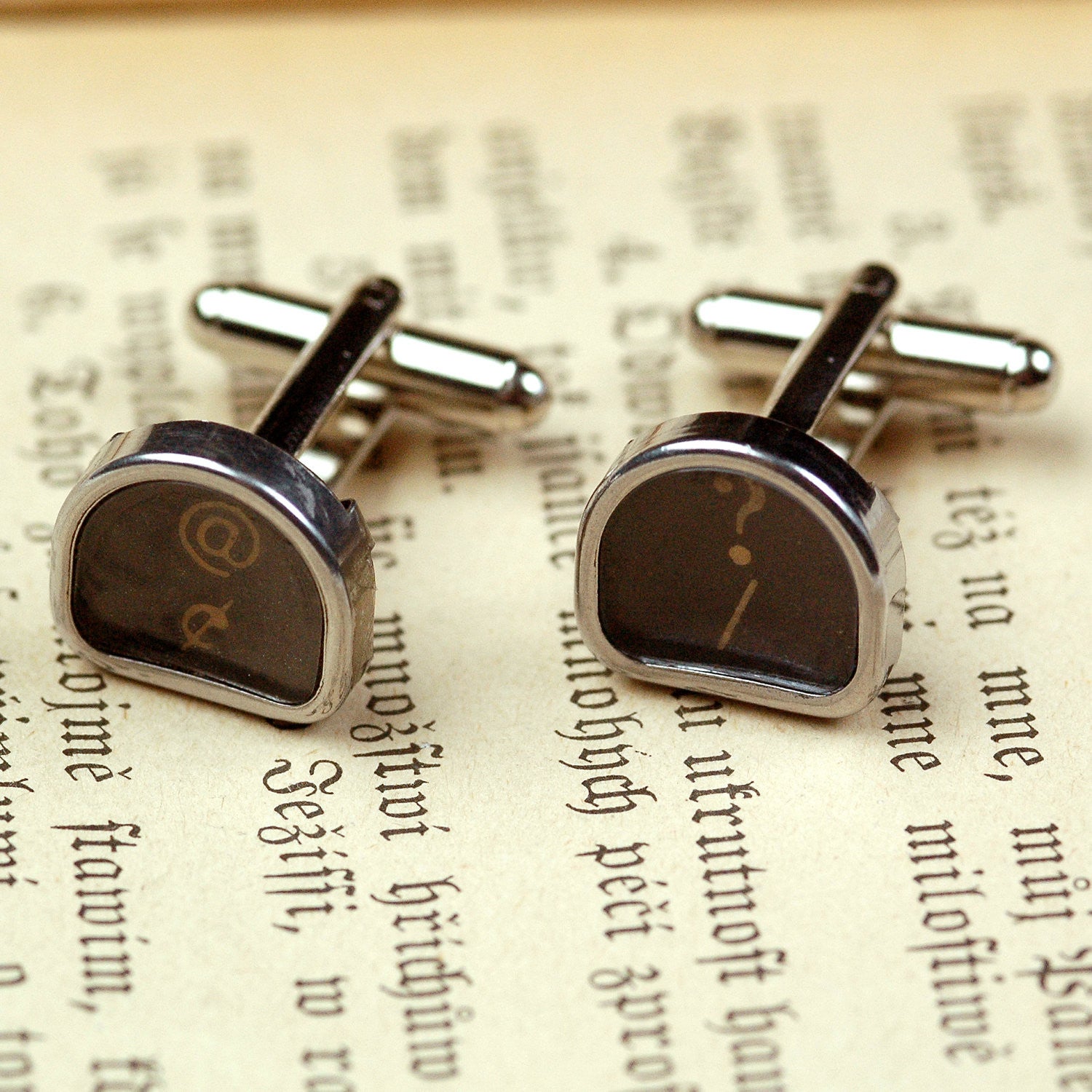 A pair of silver cufflinks with typewriter keys attached to them. Typewriter keys are metal-rimmed and tombstone-shaped with white letters on a black background.