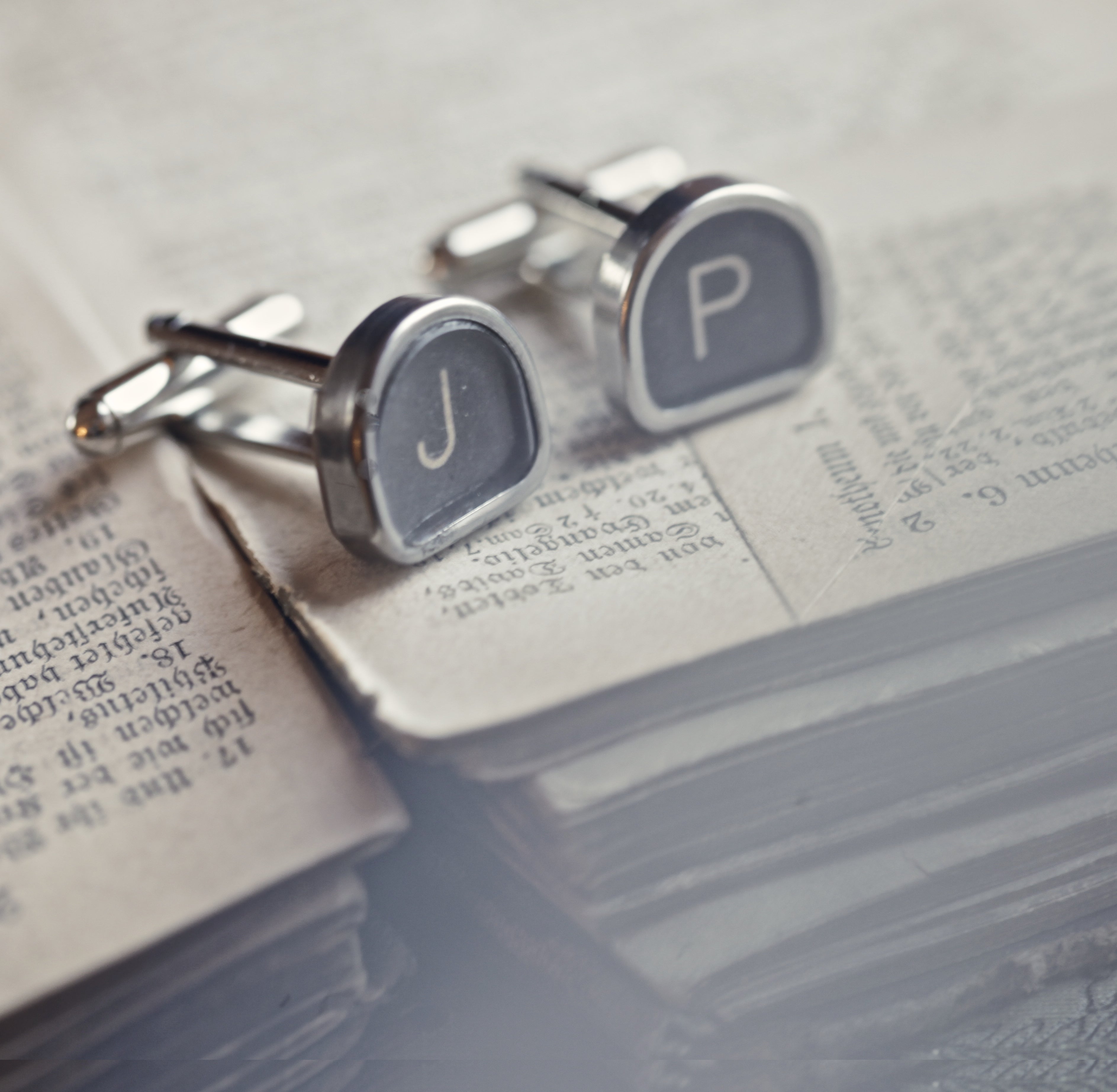 A pair of silver cufflinks with typewriter keys attached to them. Typewriter keys are metal-rimmed and tombstone-shaped with white letters on a black background.
