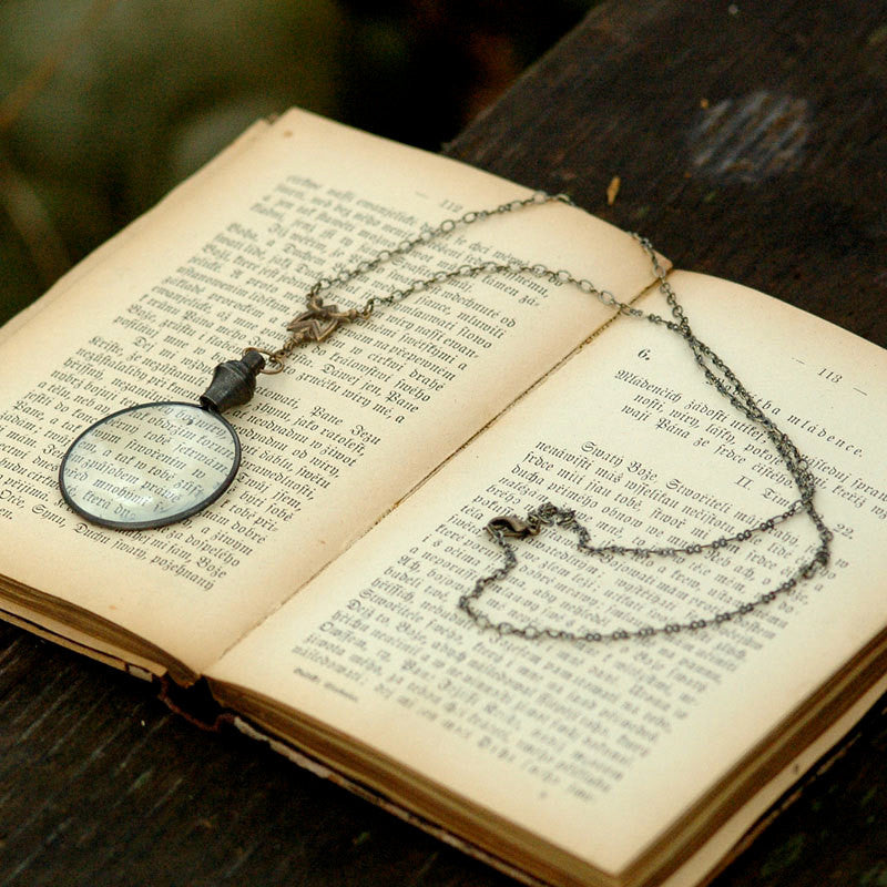 Antiqued brass round-lens glass magnifying glass attached to a chevron connector and a necklace chain.