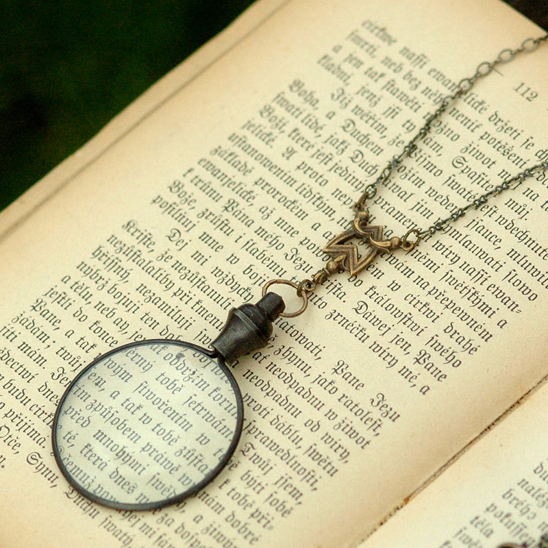 Antiqued brass round-lens glass magnifying glass attached to a chevron connector and a necklace chain.