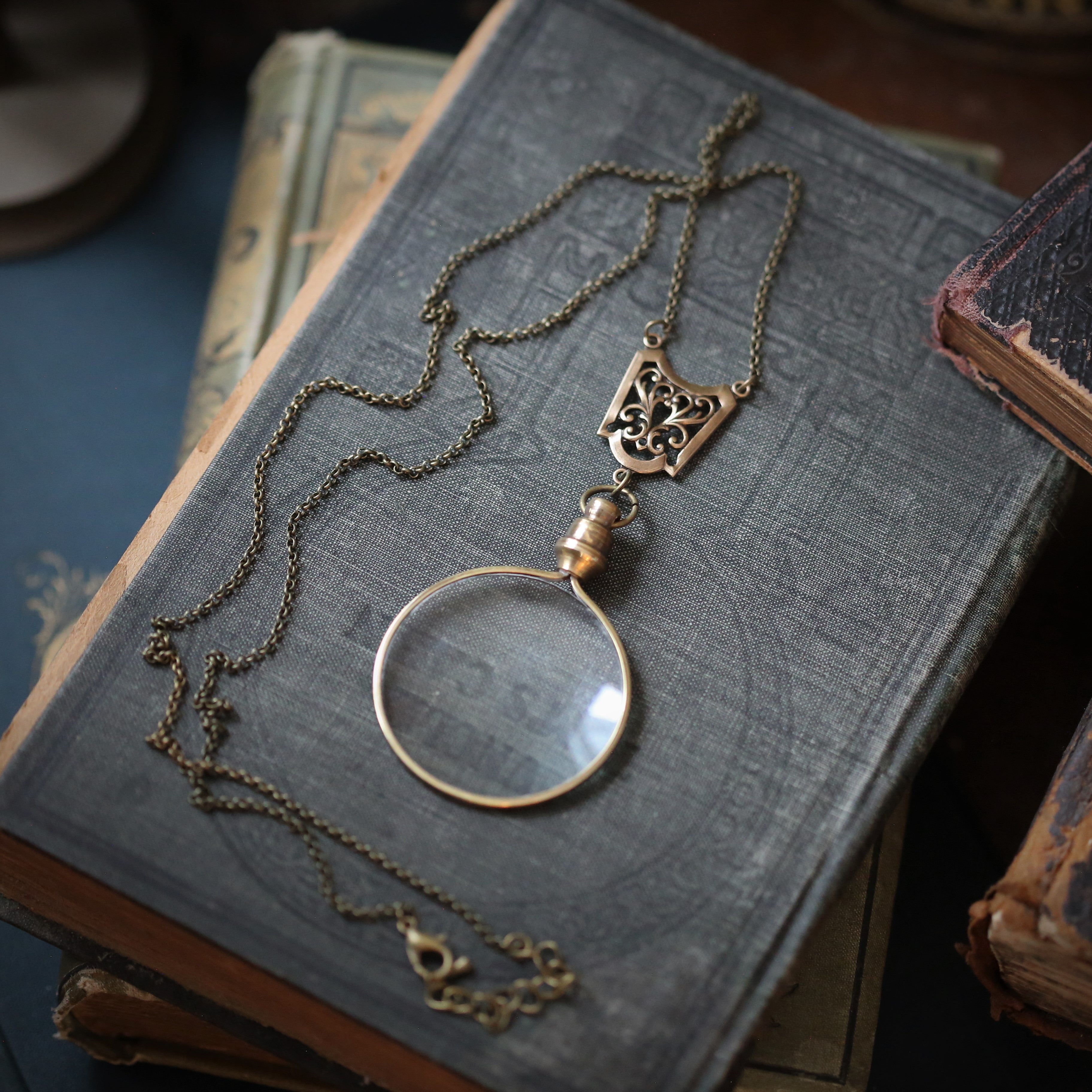 Antiqued brass round-lens glass magnifying glass attached to a shield lattice connector and a necklace chain.