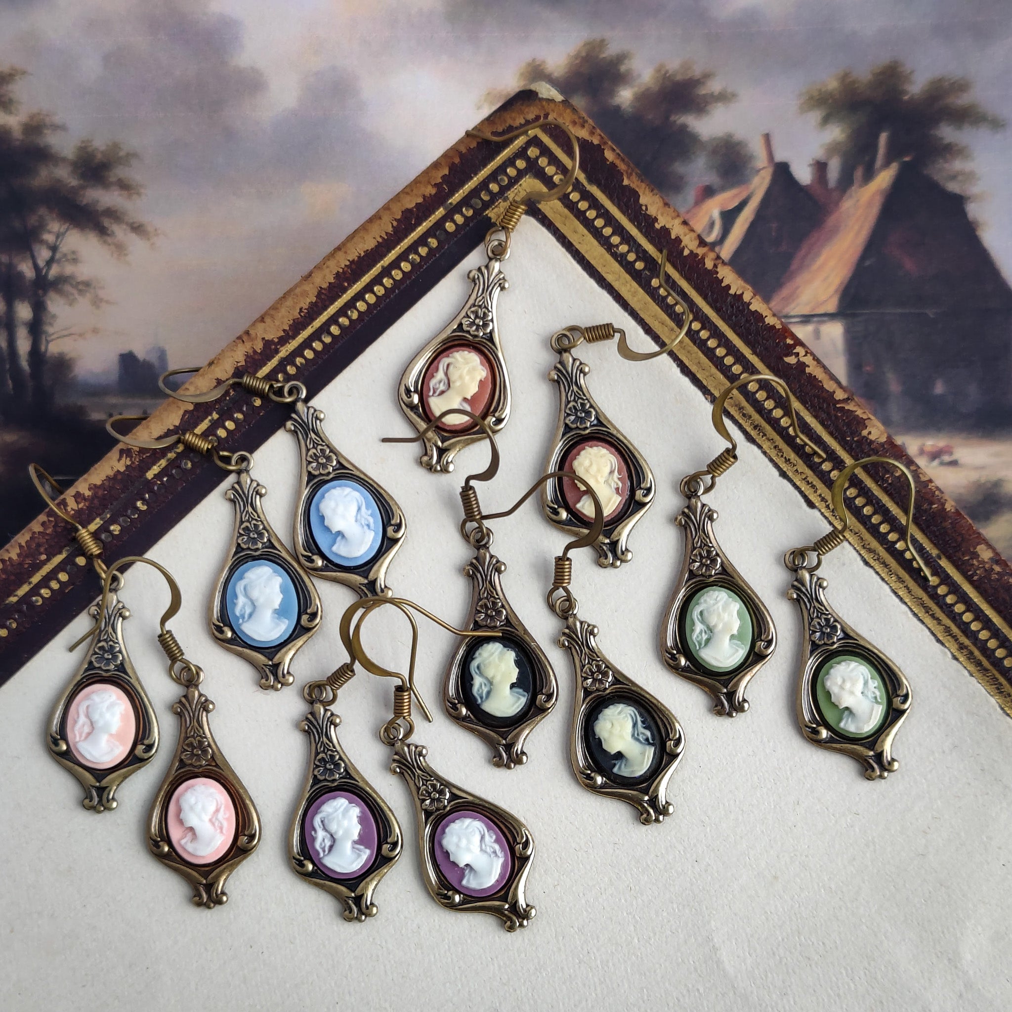 Color options for lady cameo drop earrings. Cream lady on orange,  white lady on green, white on light blue, white on pink, white on black, and white on purple. These options are on brass garden drop earring settings.