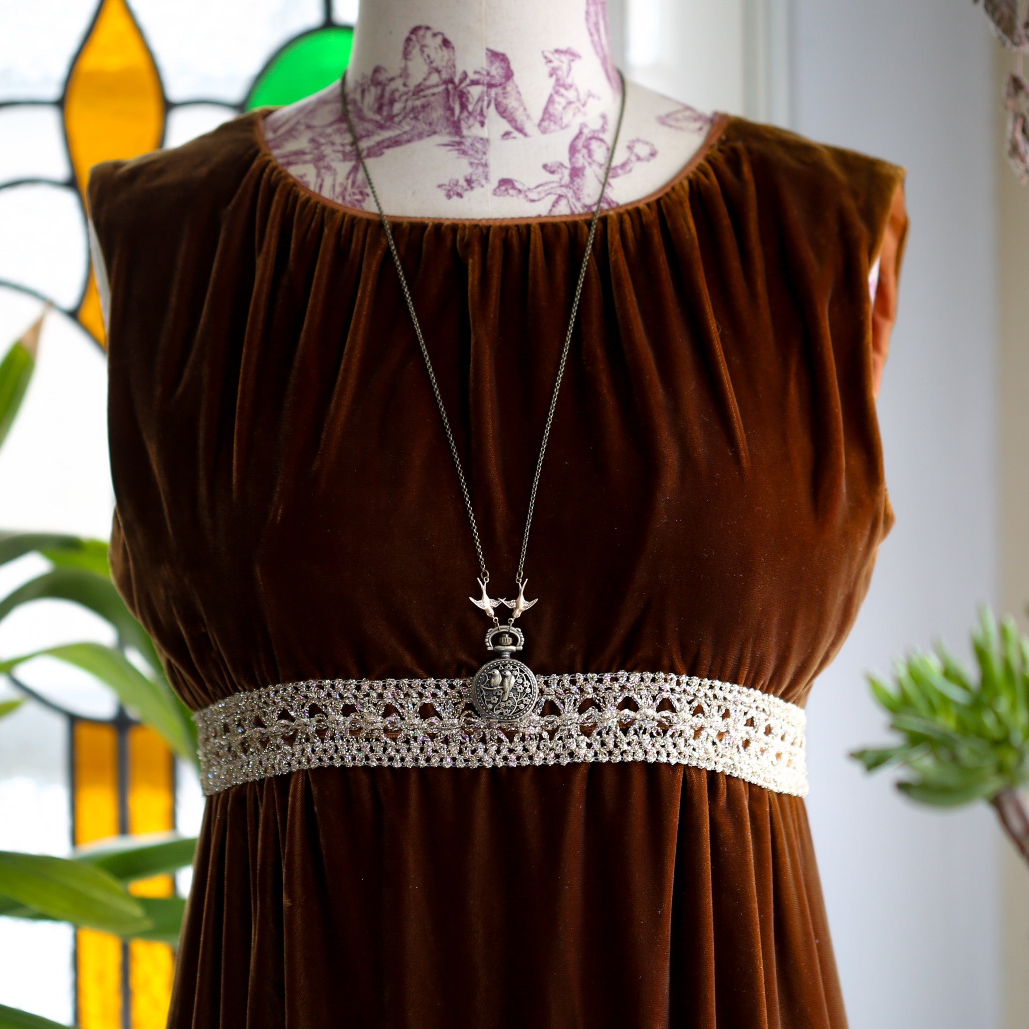 Small brass pocket watch with a cover design of two birds sitting on floral branches. Connecting the watch to the chain are two bird charms.