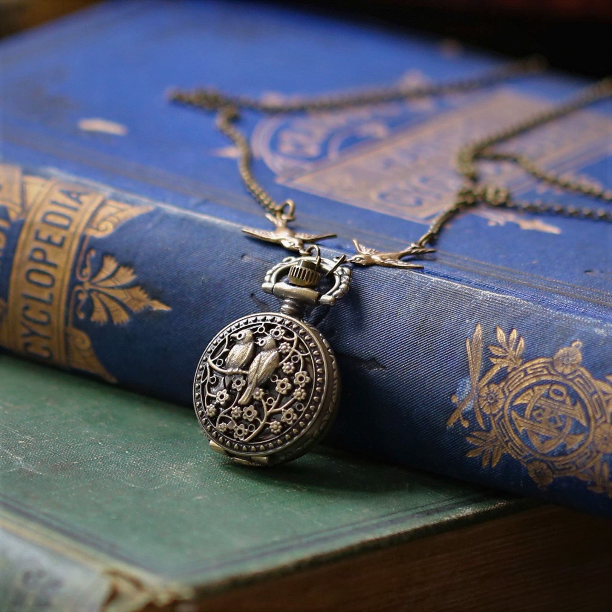 Small brass pocket watch with a cover design of two birds sitting on floral branches. Connecting the watch to the chain are two bird charms.