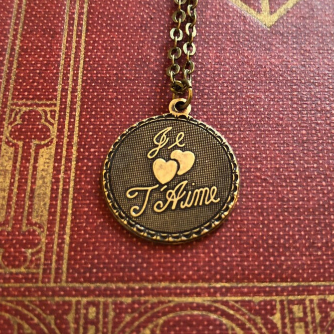 The phrase 'Je T'Aime' and two hearts on a circular brass pendant on a brass chain.