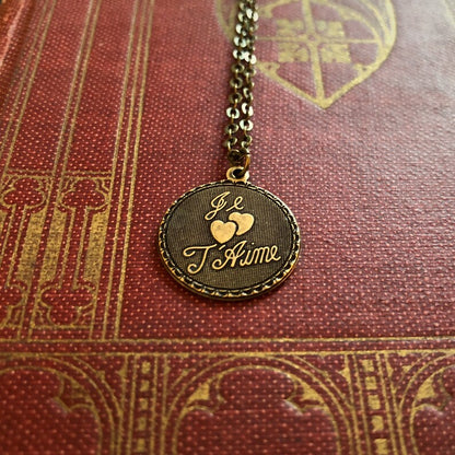 The phrase 'Je T'Aime' and two hearts on a circular brass pendant on a brass chain.