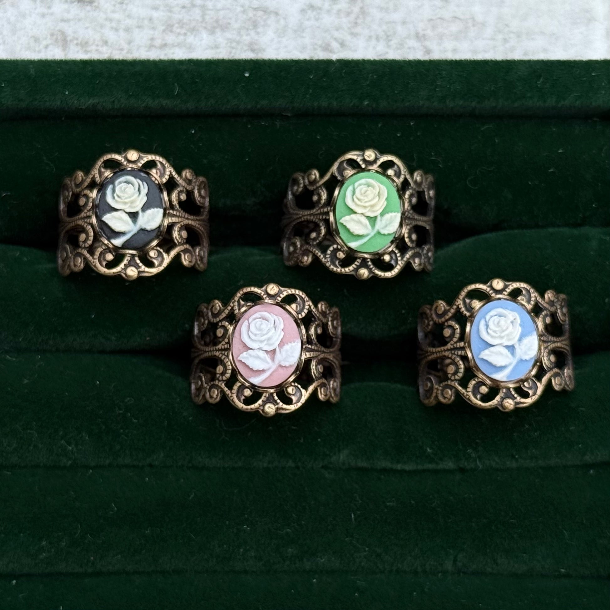 Four antiqued brass filigree rings with rose cameos. The cameos have a white flower with different colored backgrounds: black, green, pink, and blue