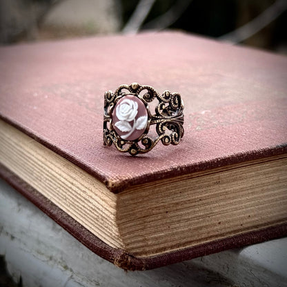 A cameo ring with a white rose on a pink background. This oval cameo is set on an adjustable brass filigree ring.