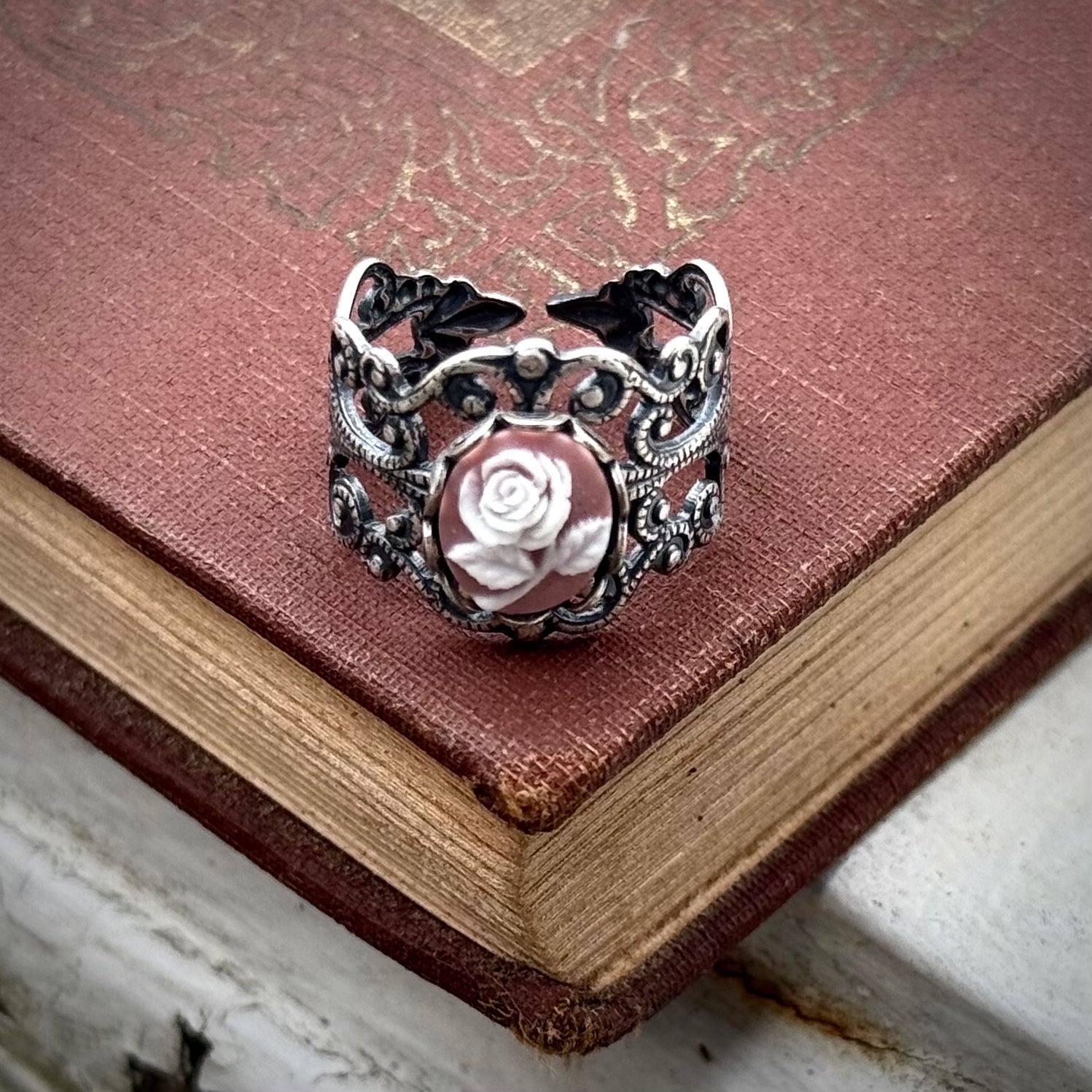 A cameo ring with a white rose on a pink background. This oval cameo is set on an adjustable silver filigree ring.