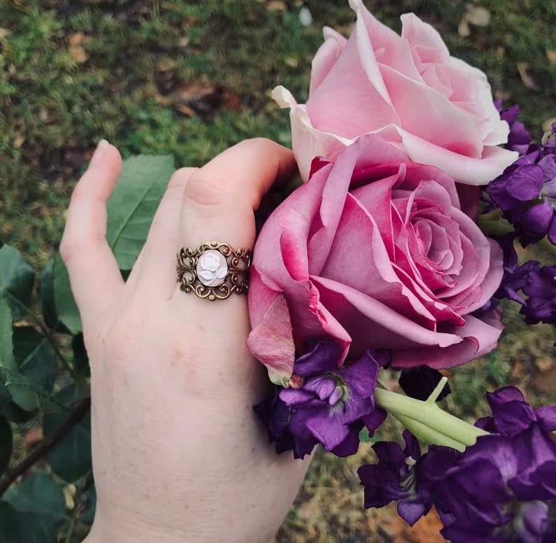 A cameo ring with a white rose on a pink background. This cameo is set on an adjustable brass filigree ring.