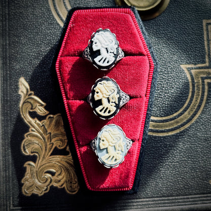 Three cameo rings: a white skeleton on black, cream skeleton on black, and a cream skeleton on a light blue background.