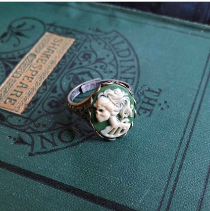 A cameo ring with a white skeleton lady on a green background. This oval cameo is set on an adjustable brass oakleaf ring.