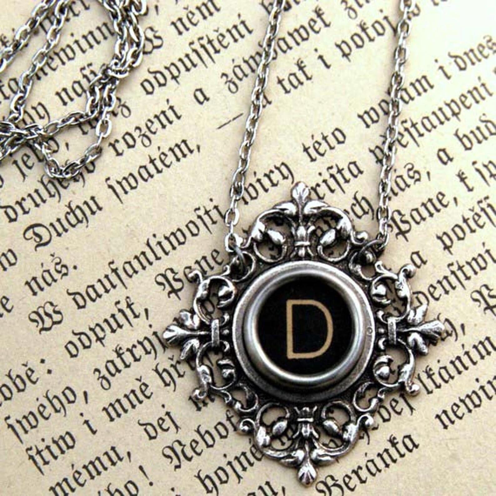 A typewriter key set on a decorative filigree pendant on a necklace chain. Typewriter key is black with an off-white colored letter D.