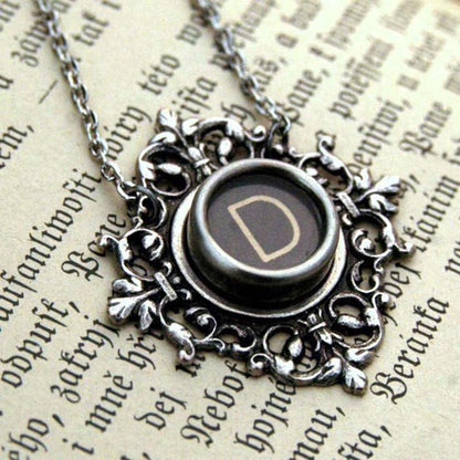 A typewriter key set on a decorative filigree pendant on a necklace chain. Typewriter key is black with an off-white colored letter D.