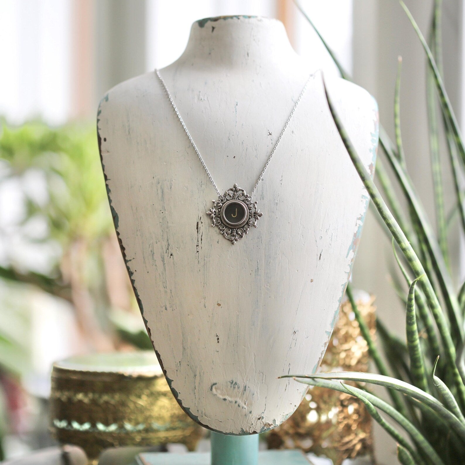 A typewriter key set on a decorative filigree pendant on a necklace chain. Typewriter key is black with an off-white colored letter J.