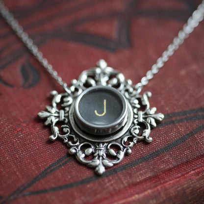 A typewriter key set on a decorative filigree pendant on a necklace chain. Typewriter key is black with an off-white colored letter J.