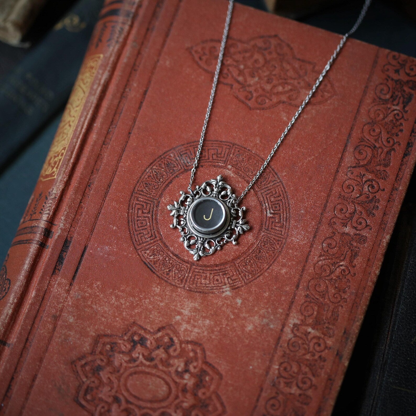A typewriter key set on a decorative filigree pendant on a necklace chain. Typewriter key is black with an off-white colored letter J.