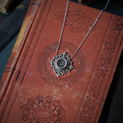 A typewriter key set on a decorative filigree pendant on a necklace chain. Typewriter key is black with an off-white colored letter J.