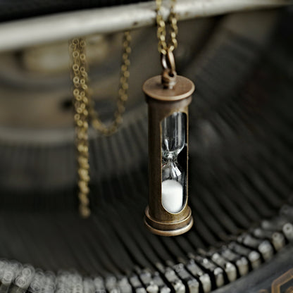 A functional hourglass in an antiqued brass frame hanging from a brass chain. The sand timer's sand is white.