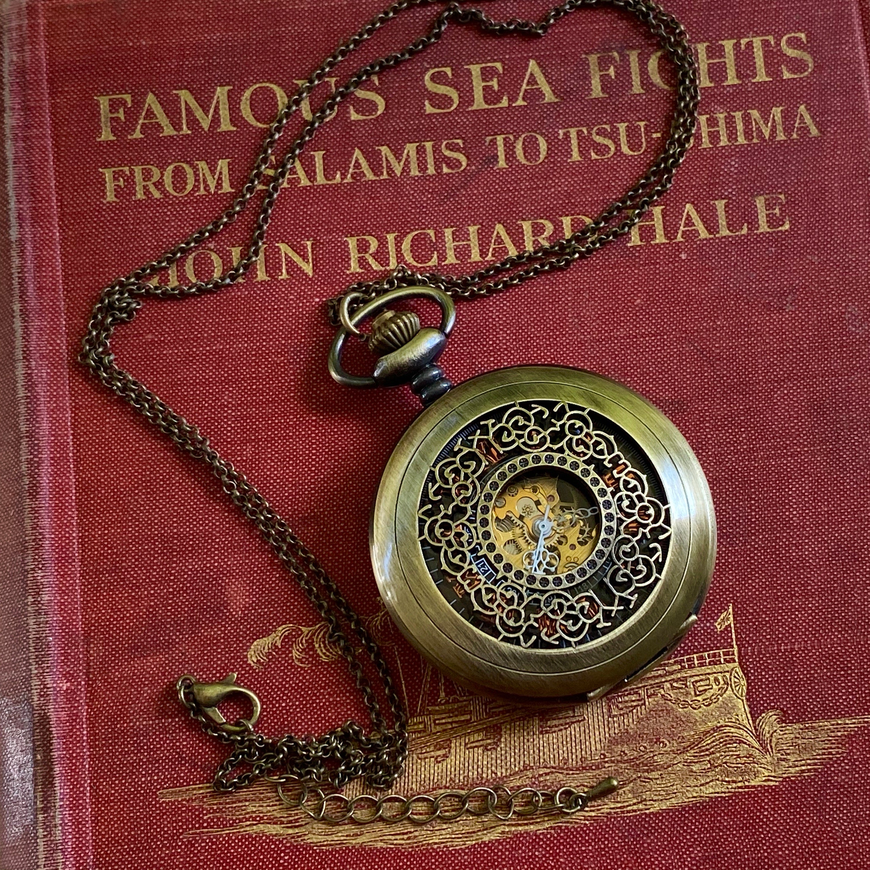 Antiqued brass mechanical pocket watch with intricate designs on the watch cover, displayed on a brass necklace chain.