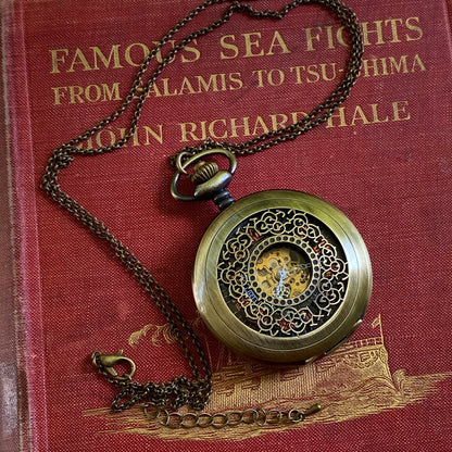Antiqued brass mechanical pocket watch with intricate designs on the watch cover, displayed on a brass necklace chain.