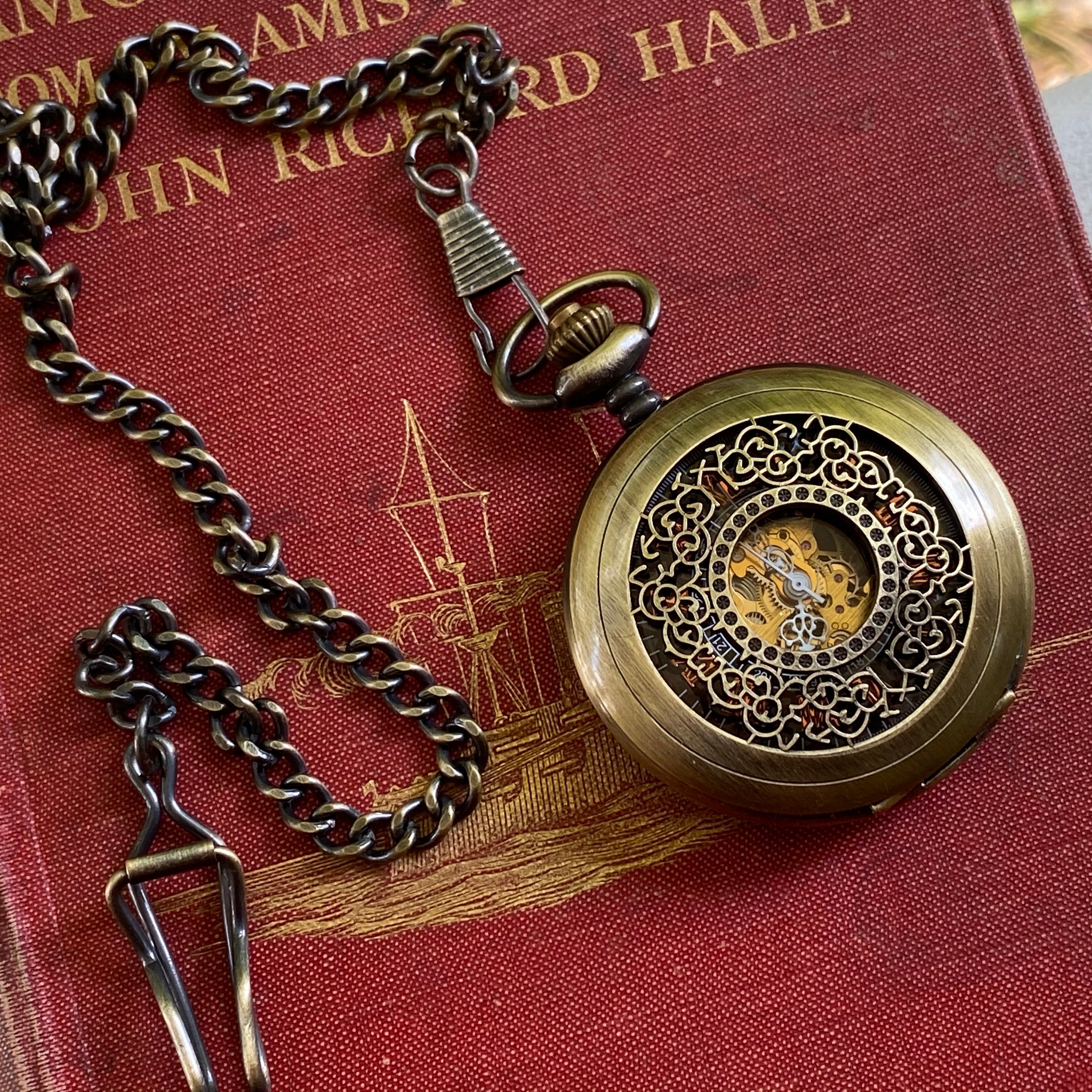 Antiqued brass mechanical pocket watch with intricate designs on the watch cover, displayed on a brass pocket chain.