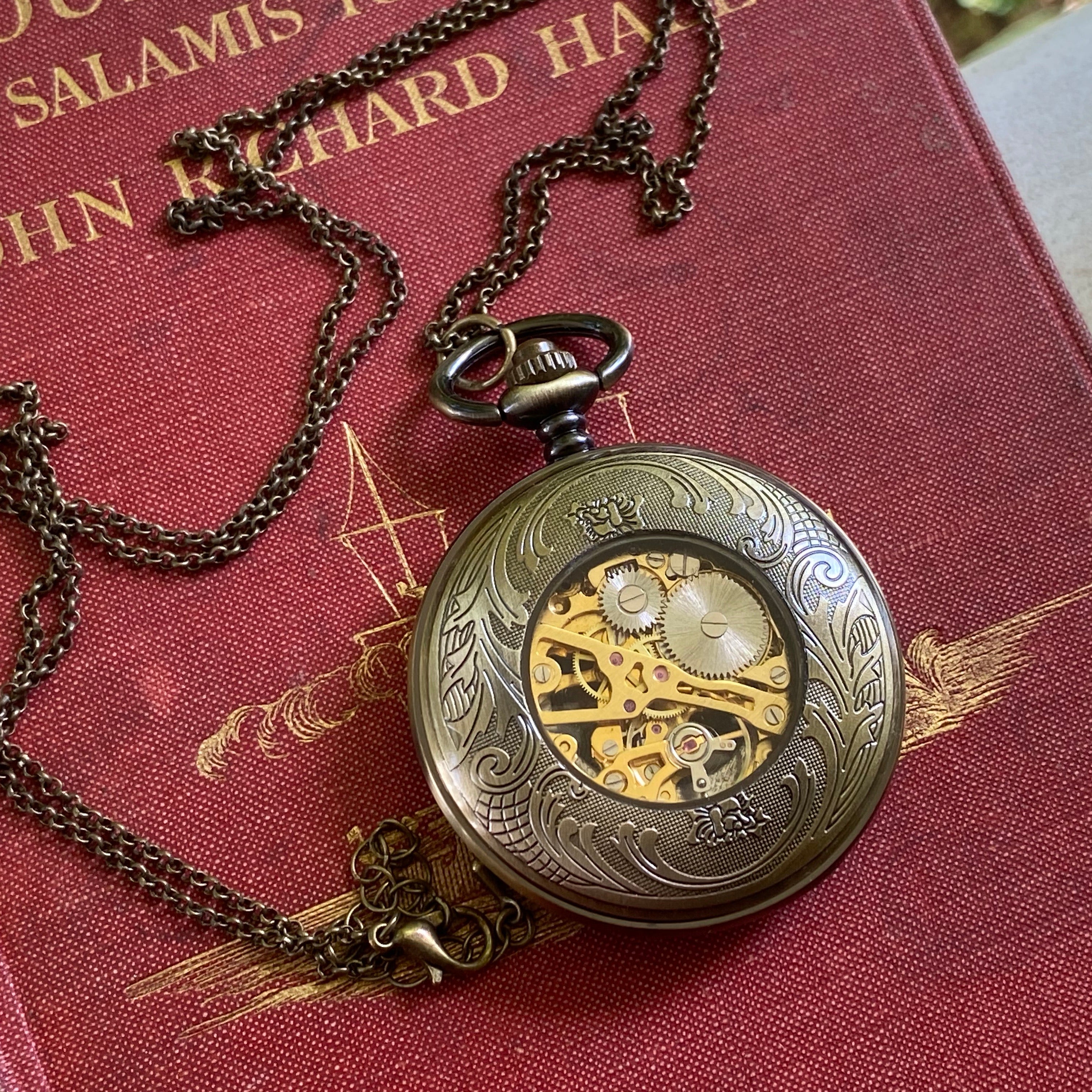 Antiqued brass Cogwheel mechanical pocket watch face down to display an ornate back panel with a window to internal gears in the center.