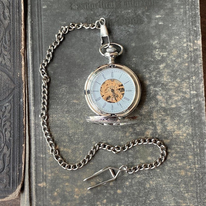 Silver mechanical pocket attached to a necklace chain. The watch is displayed open, with black roman numerals on a white boarder. Watch internal gears are visible in the center of the watch.
