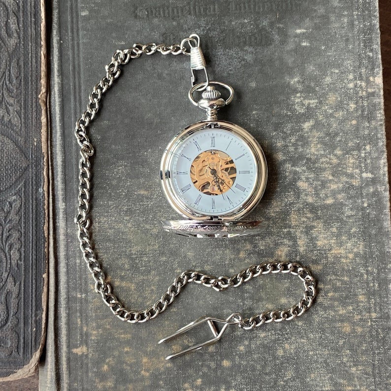 Silver mechanical pocket attached to a necklace chain. The watch is displayed open, with black roman numerals on a white boarder. Watch internal gears are visible in the center of the watch.