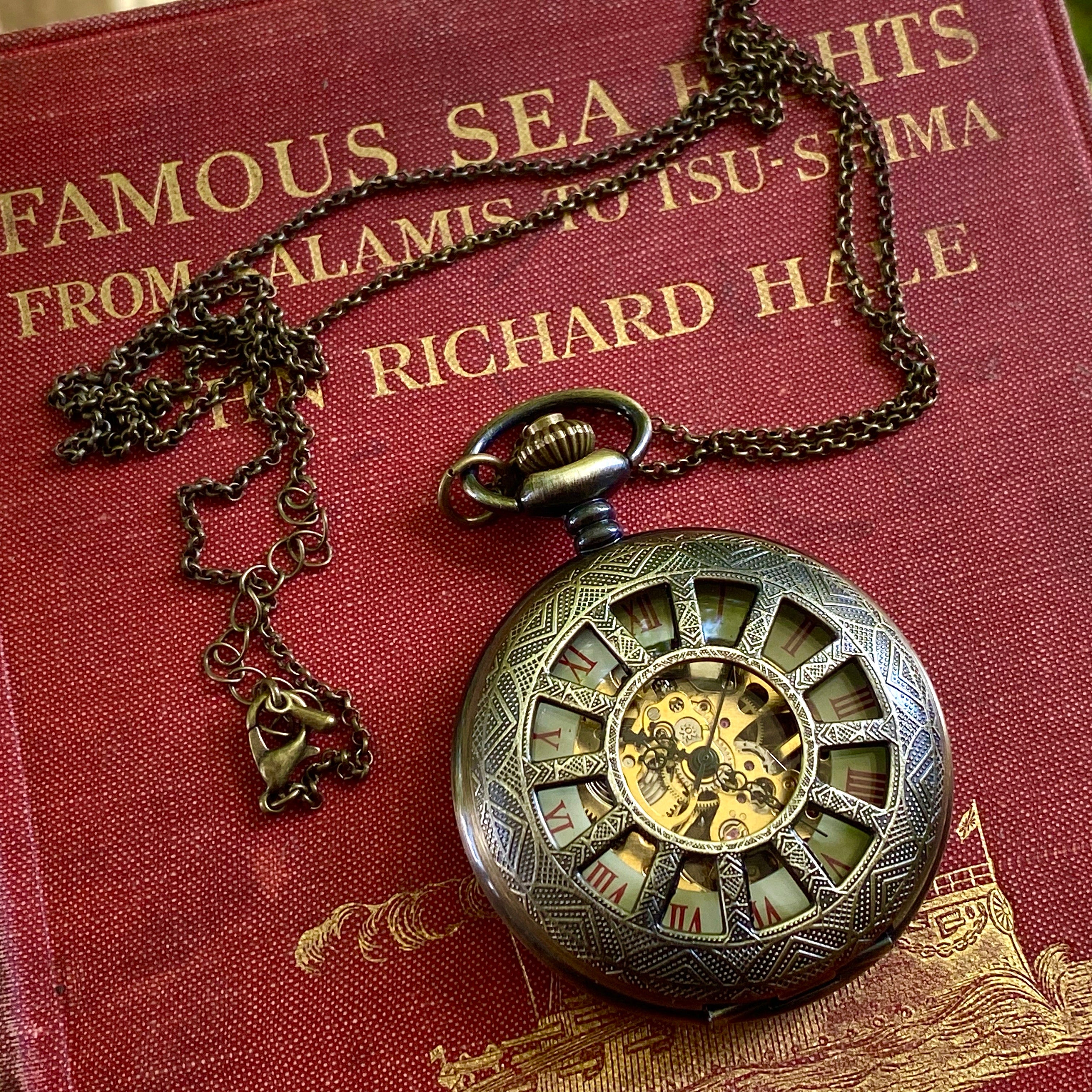Antiqued brass mechanical pocket watch with a cogwheel design on the front, attached to a brass necklace chain. The watch is closed, but internal gears are visible through the lid.