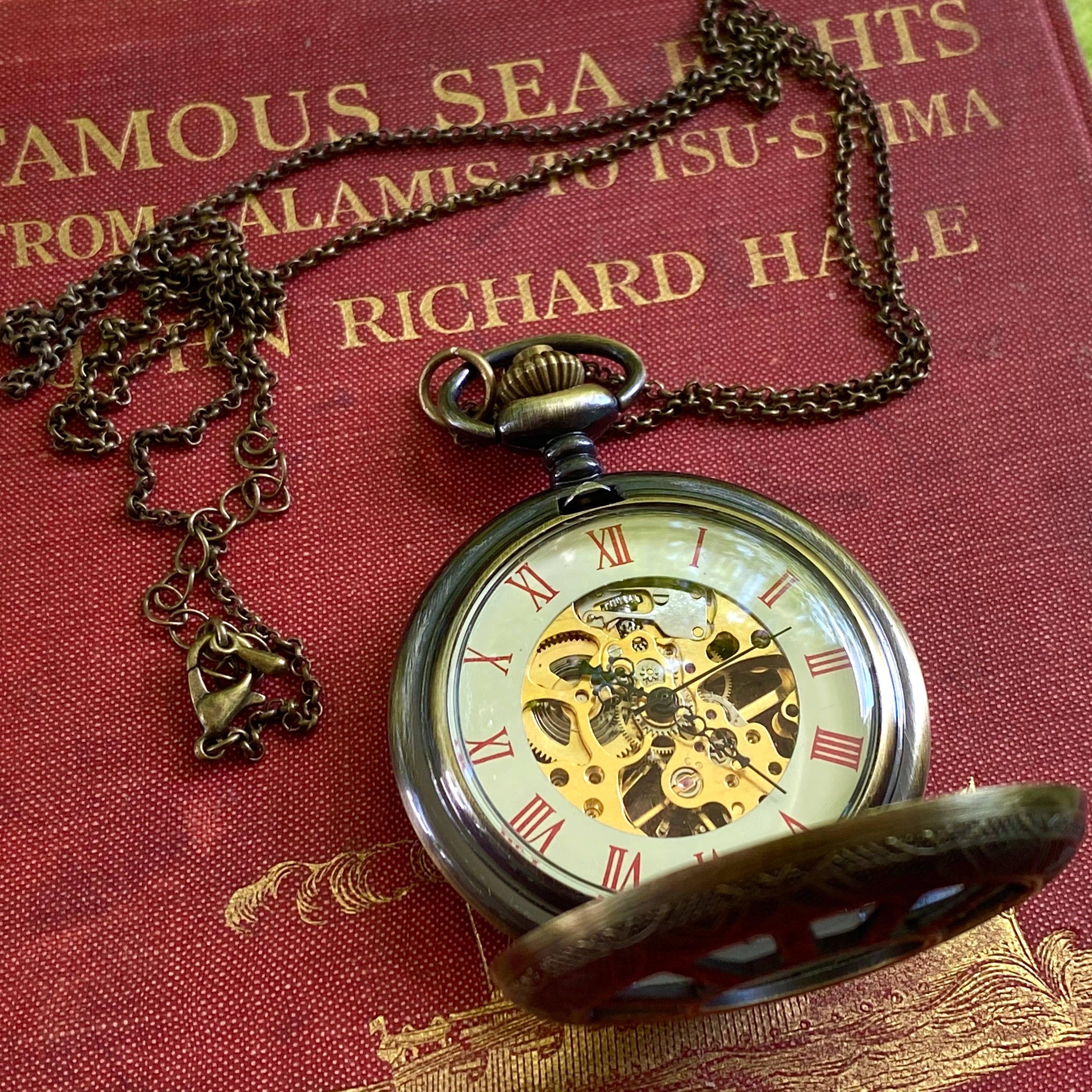 Antiqued brass mechanical pocket attached to a necklace chain. The watch is displayed open, with red roman numerals on a cream-colored boarder. Watch internal gears are visible in the center of the watch.