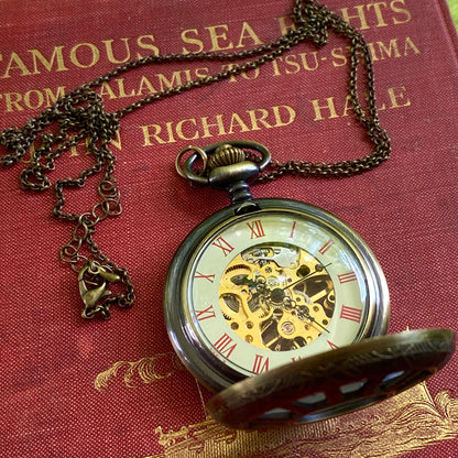 Antiqued brass mechanical pocket watch attached to a necklace chain. The watch is displayed open, with red roman numerals on a cream-colored boarder. Watch internal gears are visible in the center of the watch.