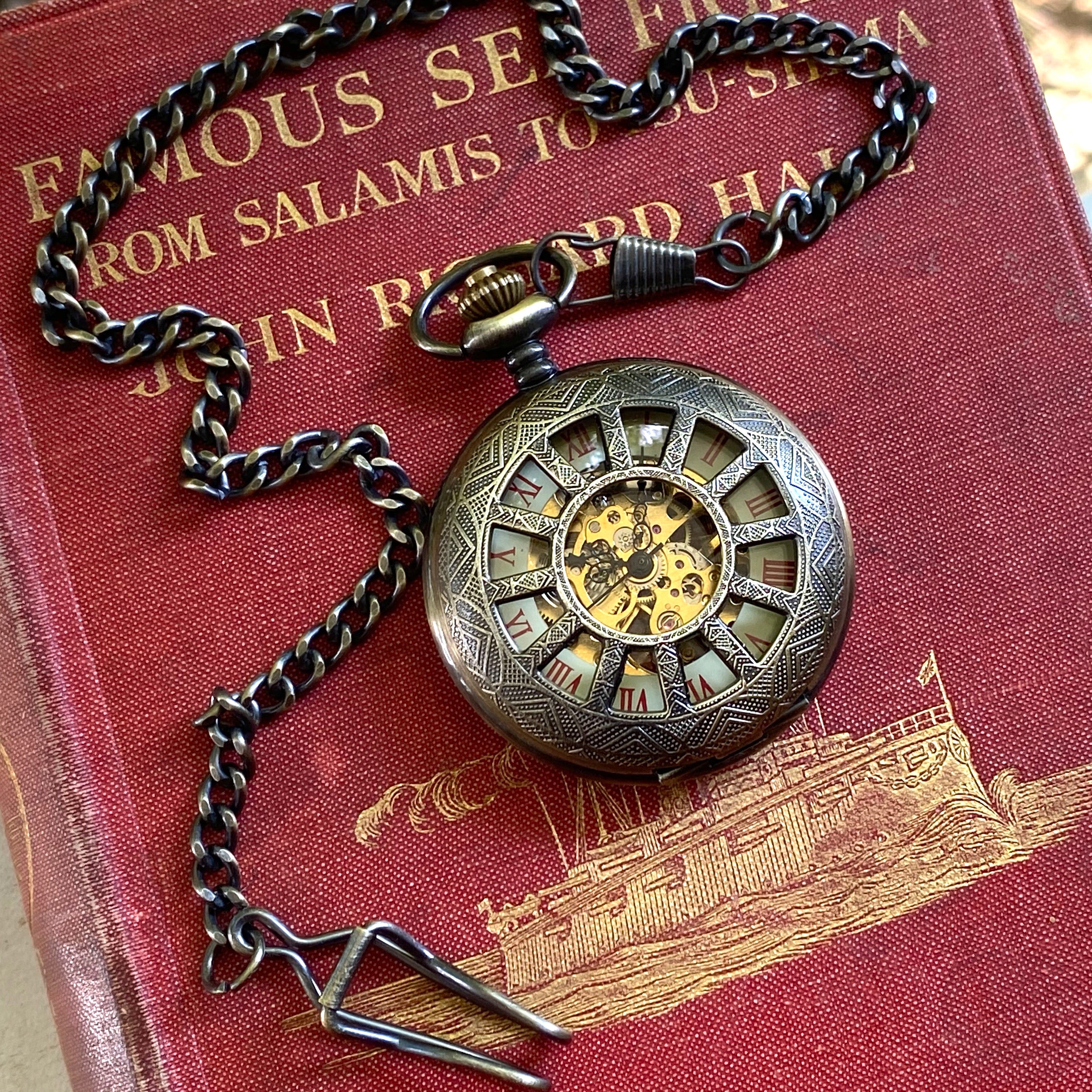 Antiqued brass mechanical pocket watch with a cogwheel design on the front, attached to a brass watch chain. The watch is closed, but internal gears and numerals are visible through the lid.