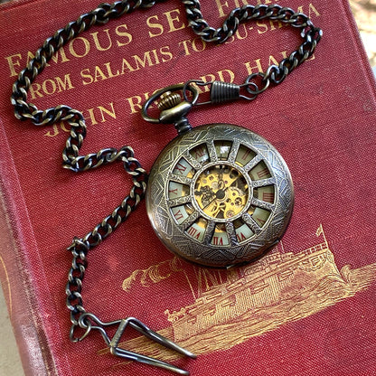 Antiqued brass mechanical pocket watch with a cogwheel design on the front, attached to a brass watch chain. The watch is closed, but internal gears and numerals are visible through the lid.