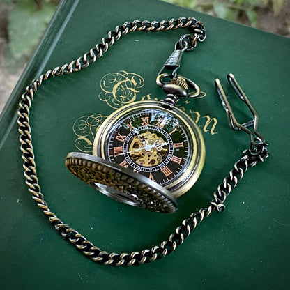Open antiqued brass mechanical pocket watch attached to a brass pocket chain. The face is black with red roman numerals and a window to the internal gears in the center.