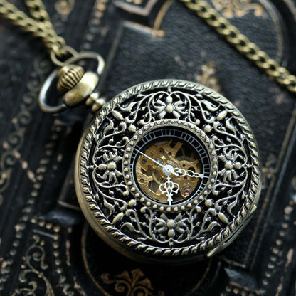 Antiqued brass mechanical pocket watch with a floral garden design attached to a brass pocket chain. The watch is closed, but internal gears are visible through the lid.