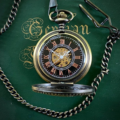 Open antiqued brass mechanical pocket watch attached to a brass pocket chain. The face is black with red roman numerals and a window to the internal gears in the center.