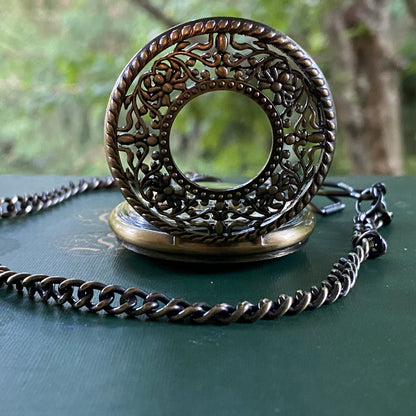 Antiqued brass mechanical pocket watch with a floral garden design attached to a brass pocket chain. The watch is closed, but internal gears are visible through the lid.