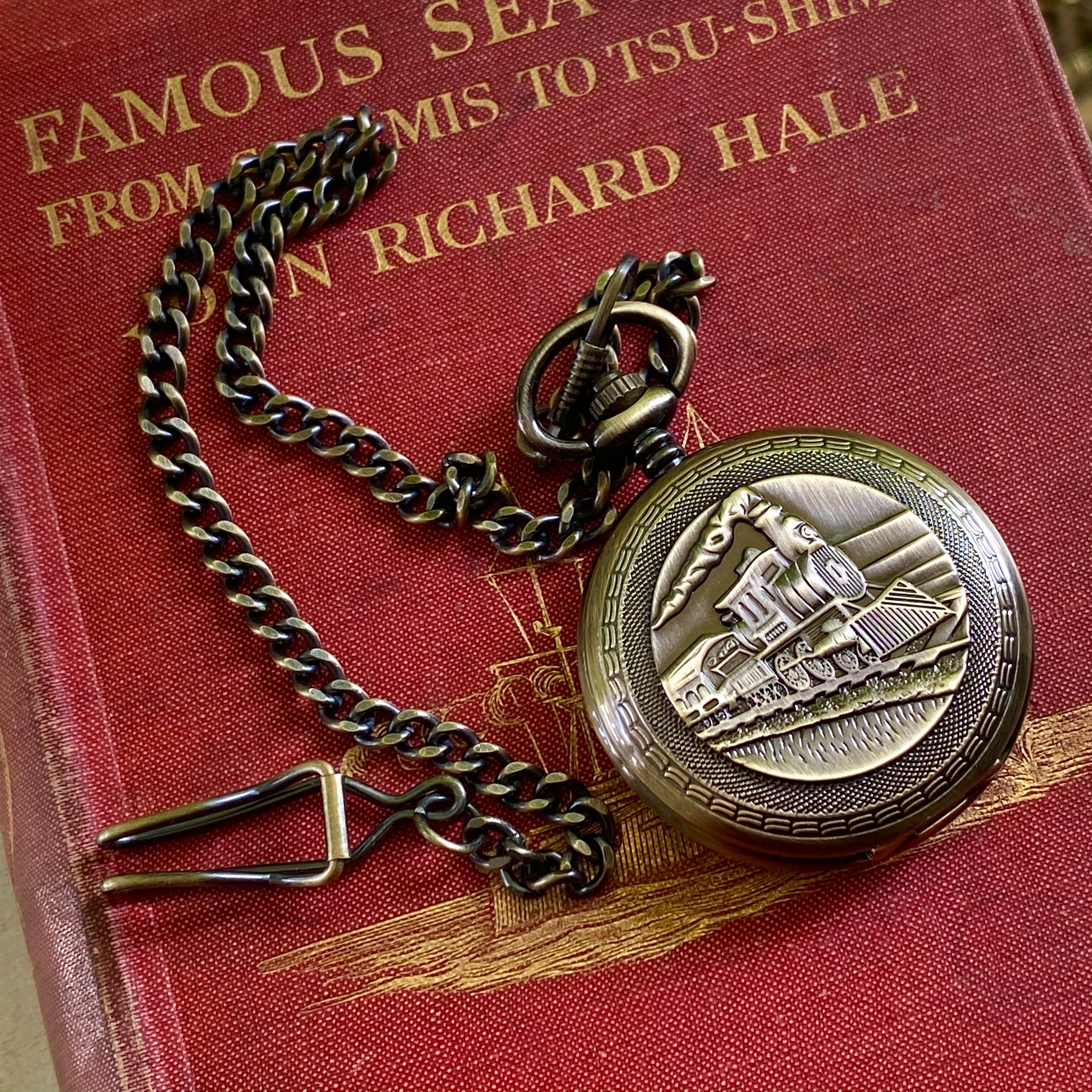 Antiqued brass mechanical pocket watch with a train design on the front, attached to a brass pocket chain.